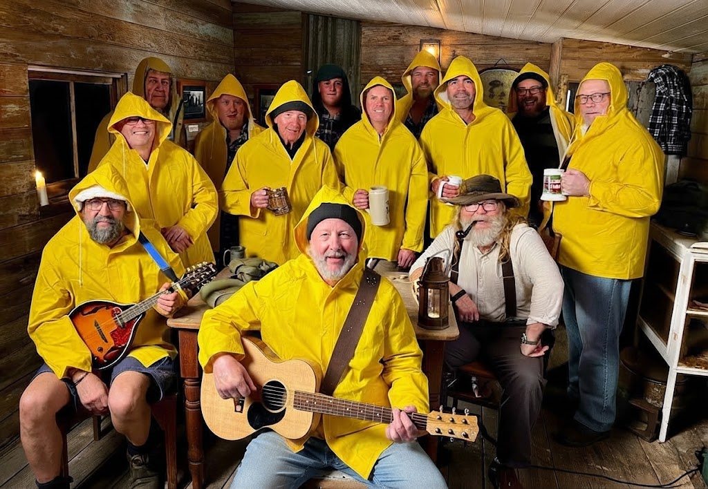 The Albany Shantymen in yellow oilskins, gathered in a timber hut with a guitar, a mandolin and mugs raised